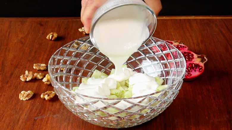 A hand is seen pouring thick yogurt from a glass bowl into a large cut-glass bowl half full of cucumber pieces. California walnuts kernels and an open pomegranate lie on the wooden counter in the background - Konkani Cucumber and Pomegranate Salad Recipe